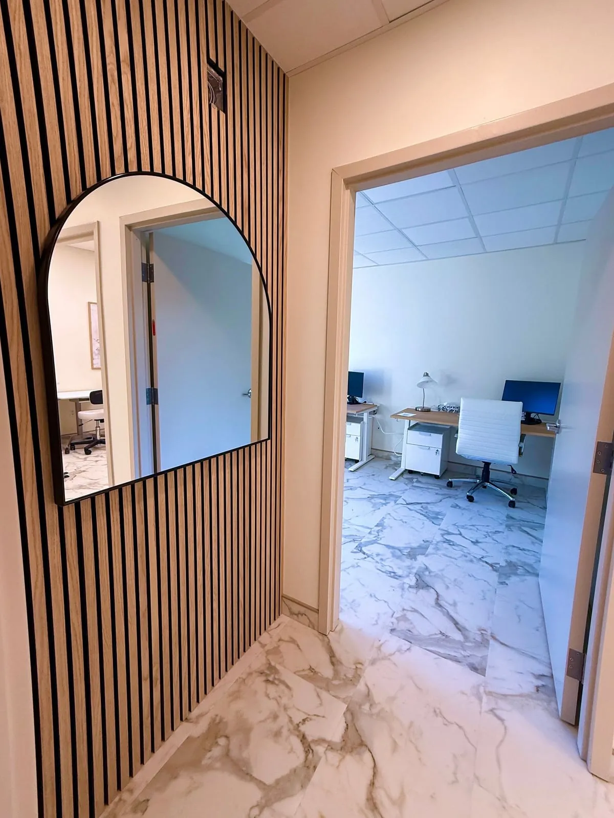 An arched mirror mounted on the slatted wood wall of the corridor, reflecting a doorway, with the physicians’ office visible through an opening further along.