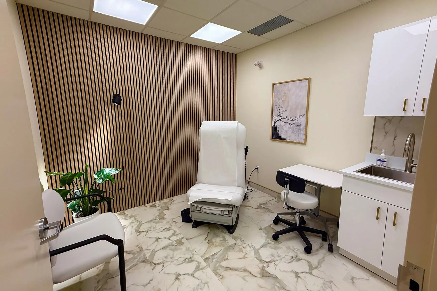An examination room: a plant against the slatted wood wall on the left, an examination chair in the centre, and a sink with cabinets on the right.