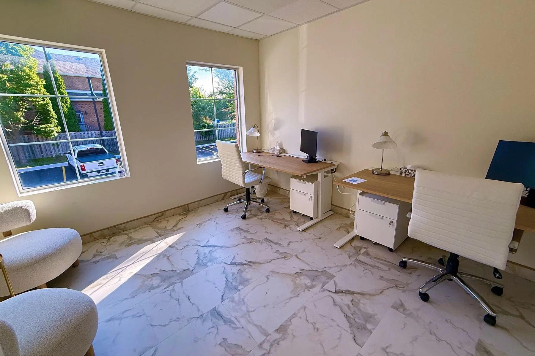 A large office with two windows, desks set along two walls, daylight falling across the floor, and two soft chairs in the near corner.